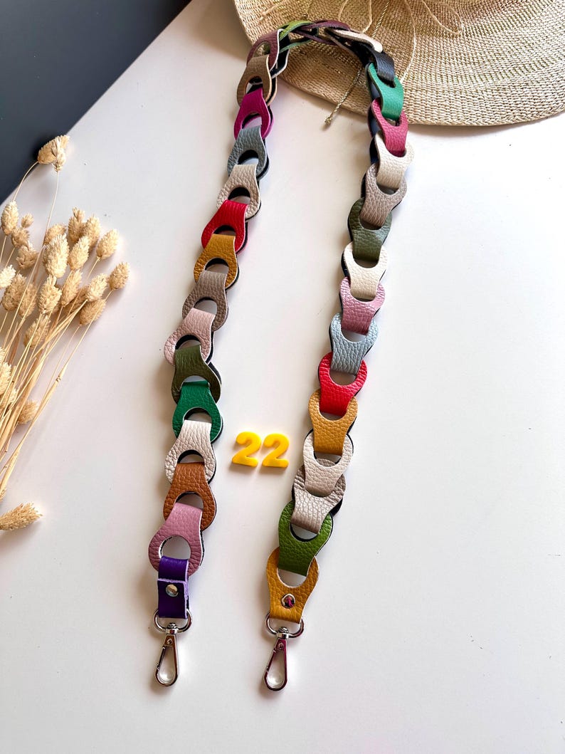 May include: A colorful handbag strap with a chain-link design. The strap is made of leather in various colors, including red, yellow, green, and purple. The strap is approximately 22 inches long and has silver-colored clasps.