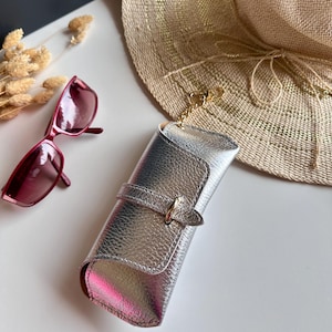 May include: Silver metallic sunglasses case with a buckle closure and a gold-colored chain. A pair of red sunglasses rests nearby, along with a straw sun hat. The case is approximately 7 inches long.
