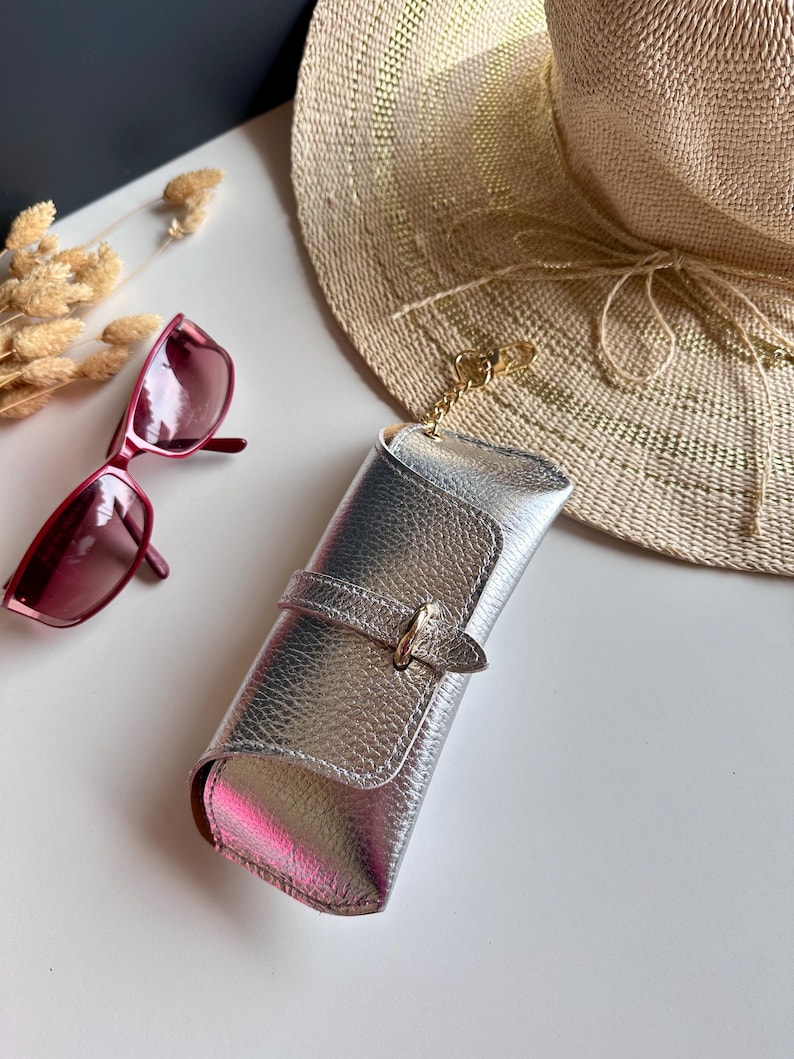 May include: Silver metallic sunglasses case with a buckle closure and a gold-colored chain. A pair of red sunglasses rests nearby, along with a straw sun hat. The case is approximately 7 inches long.
