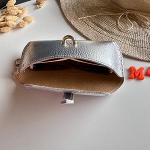 May include: Silver leatherette sunglasses case with a gold-colored ring closure. The interior is tan with a red lining. A pair of sunglasses is visible inside the case. A straw hat is in the background.