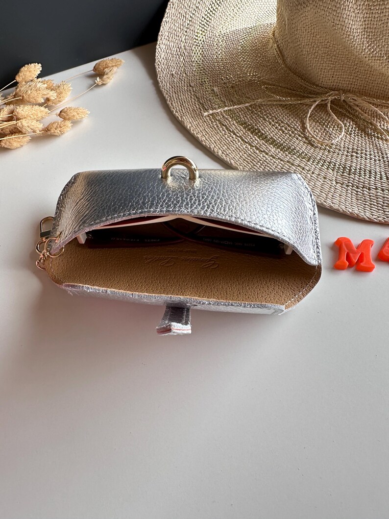 May include: Silver leatherette sunglasses case with a gold-colored ring closure. The interior is tan with a red lining. A pair of sunglasses is visible inside the case. A straw hat is in the background.