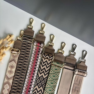 Puede incluir: Una colección de siete correas ajustables para bolsos con varios patrones tejidos en beige, marrón, negro, rojo y verde. Cada correa incluye ganchos giratorios de metal dorado y hebillas deslizantes para sujetar a bolsos de mano.