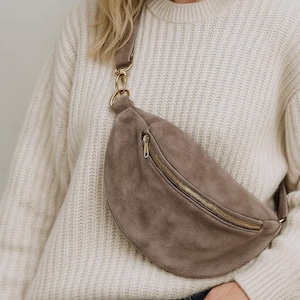 May include: A taupe suede bum bag with a gold zip and hardware. The bag is worn across the body. The person is wearing a cream-coloured jumper and blue jeans.