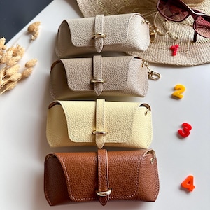 May include: Four leather eyeglass cases in beige, taupe, yellow, and brown. Each case has a gold-colored clasp and a matching strap. A straw hat and sunglasses are in the background. The numbers 1, 2, 3, and 4 are scattered around the cases.