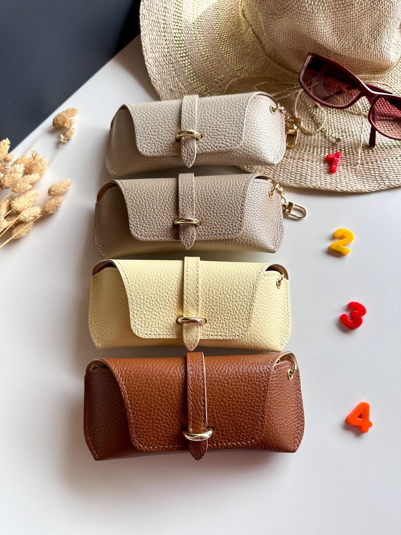 May include: Four leather eyeglass cases in beige, taupe, yellow, and brown. Each case has a gold-colored clasp and a matching strap. A straw hat and sunglasses are in the background. The numbers 1, 2, 3, and 4 are scattered around the cases.