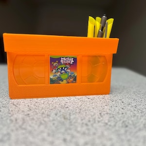 May include: Orange VHS tape-style desk organizer with a "Rugrats" label and yellow and silver pens inside.