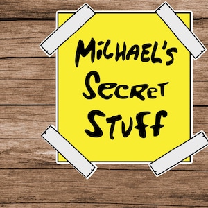 May include: A yellow sticky note with the text "Michael's Secret Stuff" in black, affixed to a wooden surface with four pieces of white tape. The note has a white border.