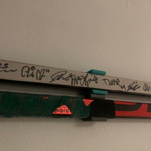 K&ouml;nnte beinhalten: Zwei Hockeyschl&auml;ger, die an einer wei&szlig;en Wand befestigt sind, mit schwarzem und gr&uuml;nem Klebeband am unteren Schl&auml;ger und einem t&uuml;rkisfarbenen und schwarzen Clip, der die Schl&auml;ger festh&auml;lt. Der obere Schl&auml;ger tr&auml;gt den Text "Shea Weber" und "Neal", der mit schwarzem Stift geschrieben wurde.