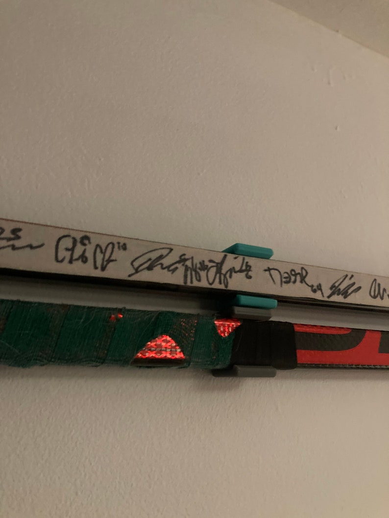 K&ouml;nnte beinhalten: Zwei Hockeyschl&auml;ger, die an einer wei&szlig;en Wand befestigt sind, mit schwarzem und gr&uuml;nem Klebeband am unteren Schl&auml;ger und einem t&uuml;rkisfarbenen und schwarzen Clip, der die Schl&auml;ger festh&auml;lt. Der obere Schl&auml;ger tr&auml;gt den Text "Shea Weber" und "Neal", der mit schwarzem Stift geschrieben wurde.