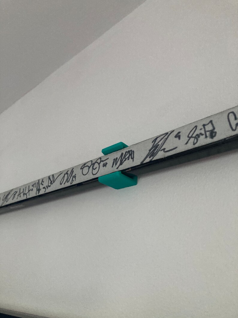 K&ouml;nnte beinhalten: Ein t&uuml;rkisfarbener Plastikhaken ist an einer wei&szlig;en Wand befestigt. Eine schwarze Metallstange mit Schriftzug ist an dem Haken befestigt.