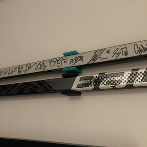 K&ouml;nnte beinhalten: Zwei Hockeyschl&auml;ger, die mit schwarzen Halterungen an einer Wand befestigt sind. Die Schl&auml;ger sind mit schwarzem Stift signiert. Der obere Schl&auml;ger tr&auml;gt die Unterschriften "AK47", "3rd", "Bliss", "88" und "Merr", der untere Schl&auml;ger die Unterschrift "Spitz".