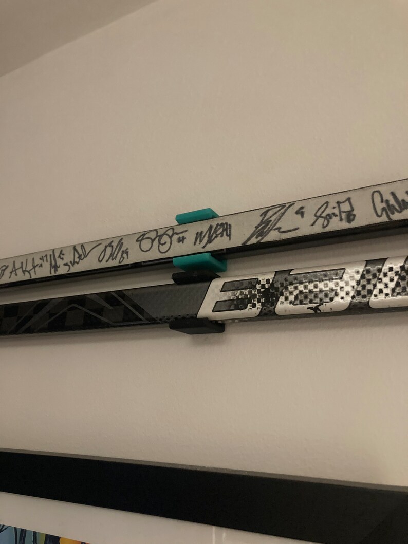 K&ouml;nnte beinhalten: Zwei Hockeyschl&auml;ger, die mit schwarzen Halterungen an einer Wand befestigt sind. Die Schl&auml;ger sind mit schwarzem Stift signiert. Der obere Schl&auml;ger tr&auml;gt die Unterschriften "AK47", "3rd", "Bliss", "88" und "Merr", der untere Schl&auml;ger die Unterschrift "Spitz".