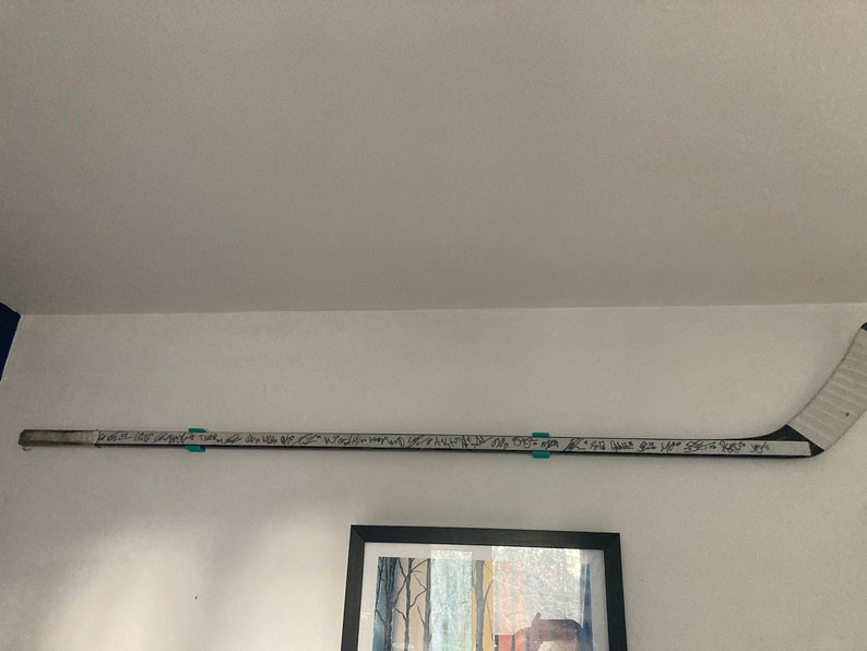 K&ouml;nnte beinhalten: Ein wei&szlig;er Hockeyschl&auml;ger mit schwarzem Klebeband auf der Klinge. Der Schl&auml;ger ist mit schwarzem Stift von mehreren Personen signiert. Der Schl&auml;ger ist an einer wei&szlig;en Wand montiert.