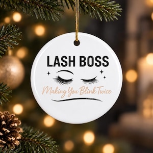May include: A round white ceramic ornament hanging by a gold string. The text LASH BOSS is printed in black above a graphic of two closed eyes with long eyelashes. Below the eyes, the text Making You Blink Twice is printed in light brown.