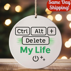 May include: A white, circular ornament with the text "Ctrl Alt + Delete My Life" in black and green. The ornament also features a power symbol and a plus sign. A string is attached for hanging.