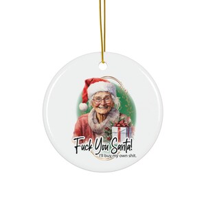 May include: A white ceramic Christmas ornament with a gold string. The ornament features a smiling elderly woman wearing a Santa hat and holding a gift. Text on the ornament reads "Fuck You Santa! I'll buy my own shit."