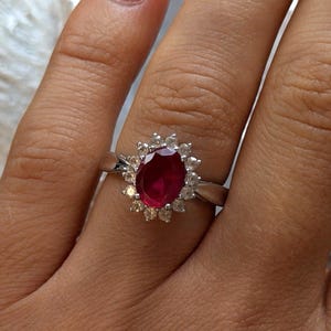Könnte beinhalten: Ein silberfarbener Ring mit einem zentralen, oval geschliffenen, tiefroten Edelstein, der von einem Kranz aus kleinen, klaren, rund geschliffenen Steinen umgeben ist. Die Ringschiene ist glatt und poliert und bildet einen Kontrast zum roten Stein.