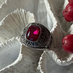 Puede incluir: Un anillo de metal plateado con una gran piedra roja oscura rectangular y facetada en el centro. La banda presenta detalles intrincados en relieve y la palabra ENGINEER es claramente visible en letras mayúsculas en el lateral.