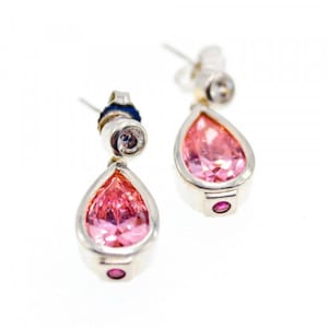 Könnte beinhalten: Ein Paar silberne Ohrhänger mit tropfenförmigen, rosa Edelsteinen. Jeder Ohrring hat einen kleinen, runden rosa Stein unten und einen kleinen, runden, klaren Stein oben. Auf weißem Hintergrund.