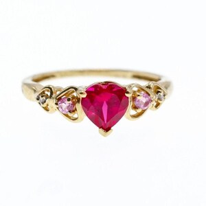 Può includere: Un anello in oro con una pietra preziosa rossa centrale a forma di cuore, affiancata da due pietre rosa rotonde più piccole e due minuscole pietre chiare, incastonate in una fascia d'oro con dettagli metallici traforati a forma di cuore.
