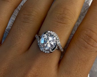 Peut inclure: Bague en argent ornée d'une grosse pierre précieuse ronde et transparente, entourée de petites pierres scintillantes. Le corps de la bague épouse gracieusement le doigt. La bague est portée sur une main aux ongles manucurés.