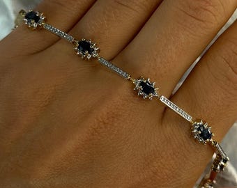 Op de afbeelding: Een goud-zilveren armband met ovale, donkerblauwe edelstenen en kleinere, heldere stenen. De armband heeft een delicaat, elegant ontwerp. De edelstenen zijn gelijkmatig verdeeld over de lengte van de armband.