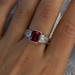 May include: A silver ring featuring a large, rectangular, deep red gemstone as the centerpiece. The band is adorned with smaller, clear gemstones. The ring is set on a hand, showcasing the jewelry's design and color.