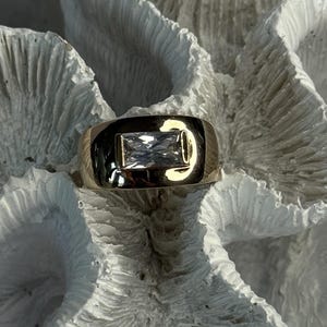 May include: A gold ring with a rectangular, clear gemstone set in a polished band. The ring is displayed on a textured, white surface, creating a contrast between the shiny metal and the matte background. The gemstone reflects light, adding sparkle.