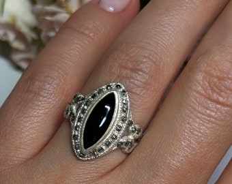 May include: A silver ring with a black oval gemstone surrounded by smaller stones. The ring is on a finger.