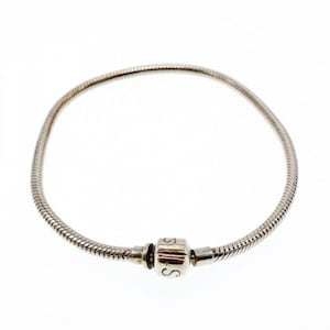 May include: A silver-toned bracelet with a snake chain design. The bracelet features a cylindrical clasp with engraved details. The clasp is connected to the chain with small metal connectors. The bracelet is displayed against a white background.