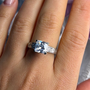 May include: A silver ring with a large, square, clear gemstone. The band features an ornate, engraved design. The ring is shown on a person's finger, highlighting the intricate details and the brilliance of the stone.