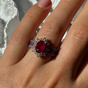 May include: A silver ring featuring a large oval ruby center stone surrounded by smaller white stones. The ring has a halo design and is flanked by rows of smaller red stones. The ring is on a person's finger.