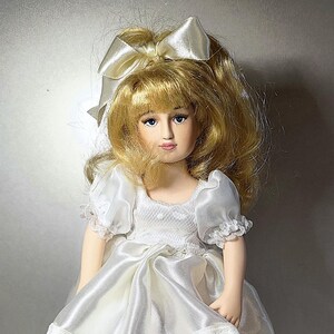 Peut inclure: Une poupée en porcelaine portant une robe en satin blanc avec une bordure en dentelle et des chaussures blanches. La poupée est sur un socle de boîte à musique en velours rouge.