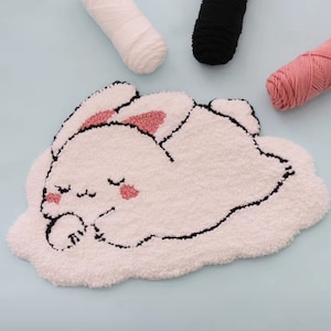May include: A white fluffy rug with a black outline of a sleeping bunny with pink ears and cheeks. The rug is on a light blue background with balls of yarn in white, black, and pink.