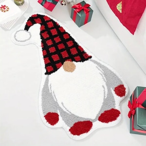 Op de afbeelding: Een kaboutervormig tapijt met een witte baard en een rood en zwart geruite hoed. Het tapijt heeft grijze armen en rode voeten. Dit feestelijke tapijt is perfect voor de feestdagen.
