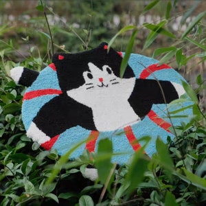 May include: A round, blue and red striped rug with a black and white cat design. The cat has a white face with black eyes and a red nose.
