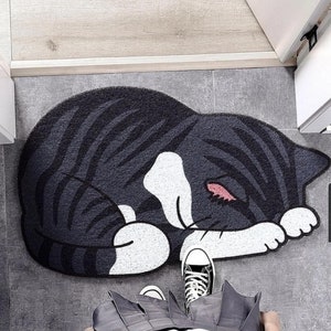 May include: A black and white cat-shaped doormat featuring a sleeping cat design. The cat is curled up with its paws tucked under its chin. The doormat is made from a soft, plush material.