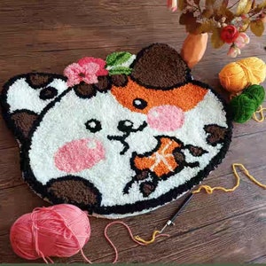 Peut inclure: Un tapis fait à la main en forme de visage de chat, avec des sections blanches, marron, orange et roses, avec des détails noirs. Une pelote de fil rose et une pelote de fil jaune sont à proximité, suggérant un projet artisanal.