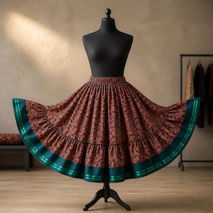 May include: A full-length skirt featuring a dark red and black floral pattern with a wide, solid dark teal hem. The hem is accented with two horizontal green ribbon stripes. The skirt has a gathered waist and a flared, voluminous silhouette.