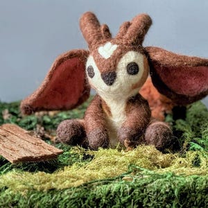 May include: A small, brown and white felt fawn with large ears sits on a bed of green moss. The fawn has a white heart-shaped marking on its forehead and large, black eyes.