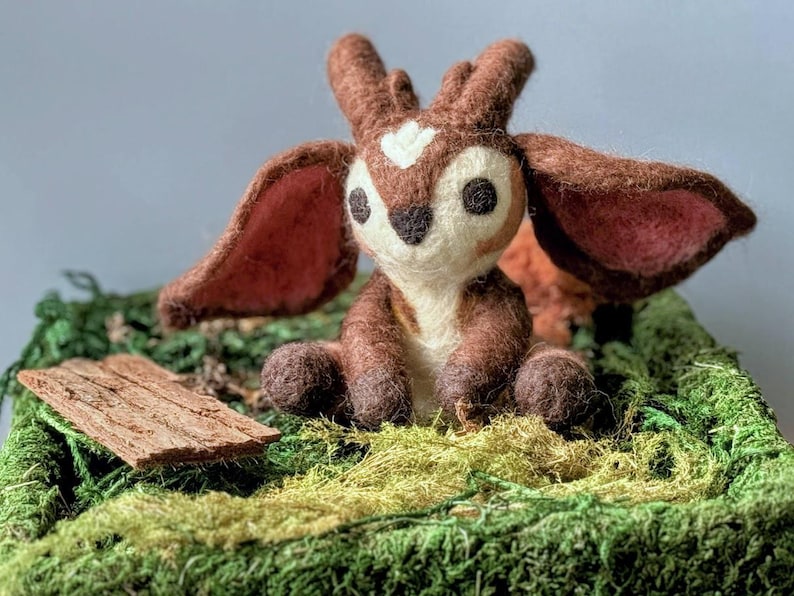May include: A small, brown and white felt fawn with large ears sits on a bed of green moss. The fawn has a white heart-shaped marking on its forehead and large, black eyes.
