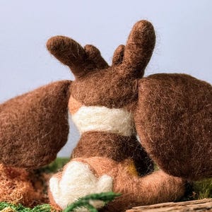 May include: A brown and white felt animal toy with large ears and a white belly. The toy is sitting on a bed of green moss.