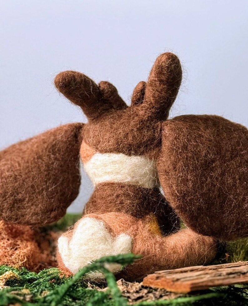 May include: A brown and white felt animal toy with large ears and a white belly. The toy is sitting on a bed of green moss.