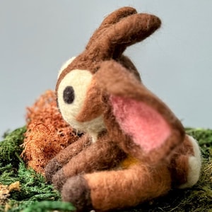 May include: A brown and white needle felted deer figurine with pink ears, sitting on a bed of green moss and brown twigs.