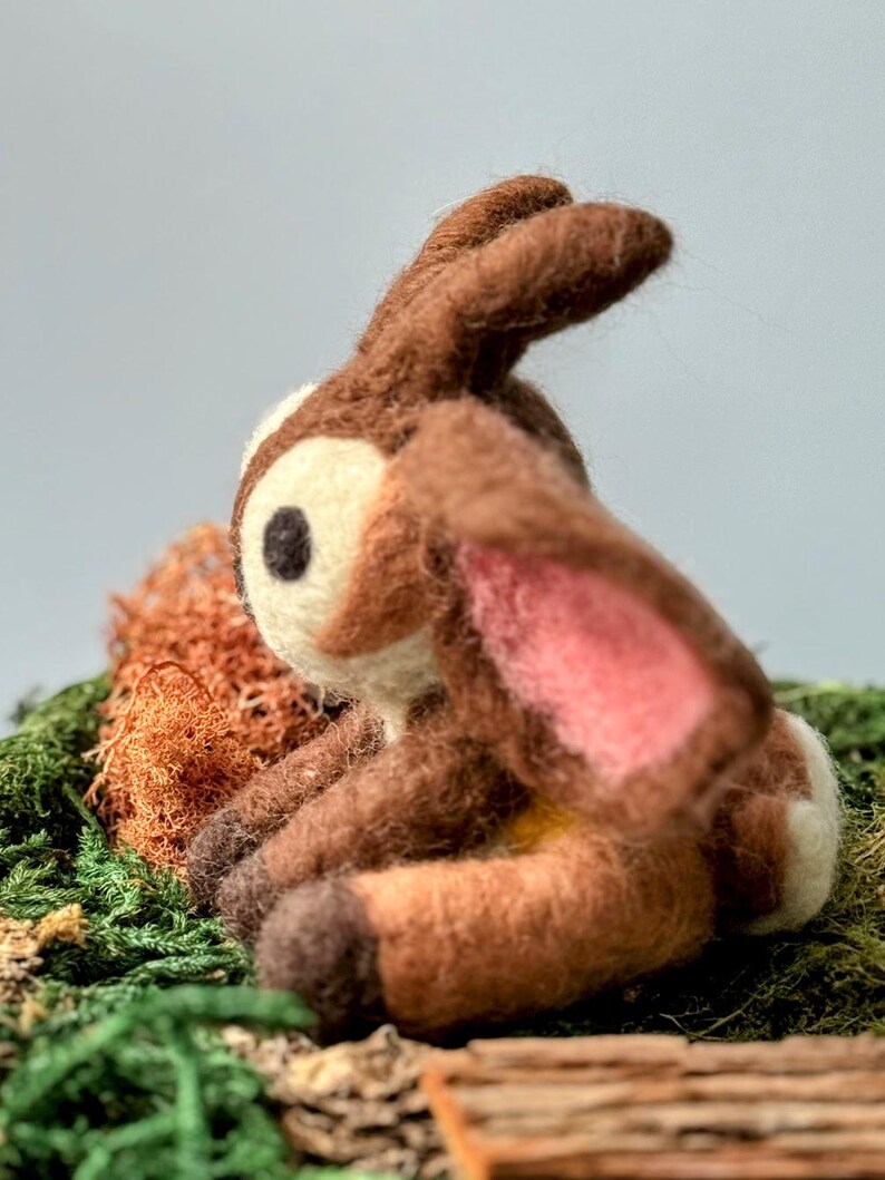 May include: A brown and white needle felted deer figurine with pink ears, sitting on a bed of green moss and brown twigs.