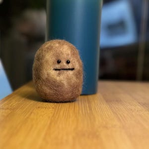 May include: A brown felt ball with a simple face, featuring two black eyes and a black mouth, is sitting on a wooden surface.