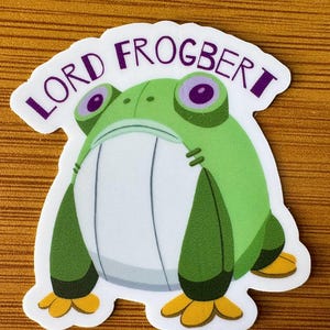 May include: A sticker featuring a cartoon frog with the text "LORD FROGBERT" in purple. The frog is green with large purple eyes, a white belly, and yellow feet. The sticker has a white border.