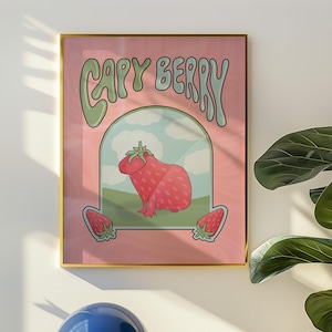 A framed poster with a pink background with green and light blue text that reads 'Capy Berry'. The poster features a cartoon capybara with a strawberry on its head. The capybara is standing on a green patch of grass with a blue sky and white clouds in the background. Two strawberries are on either side of the capybara.