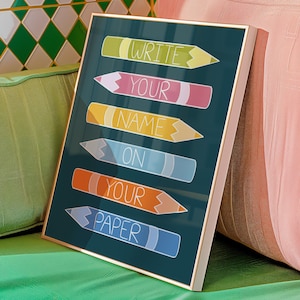 May include: A framed art print with a dark teal background. The print features colourful pencil illustrations with the words "WRITE YOUR NAME ON YOUR PAPER" in white text. The frame is gold.