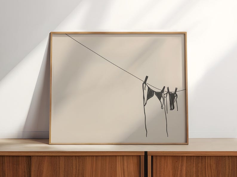 Horizontal art print of a bikini set hanging on a clothesline. The bathing suit is dark grey and in the bottom right of the artwork. The background is tan/beige.